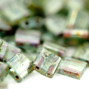 May include: A collection of rectangular glass beads featuring a translucent light green base with iridescent metallic finishes and internal streaks of reddish-brown and gold. Each bead has two small holes drilled through the center for threading.