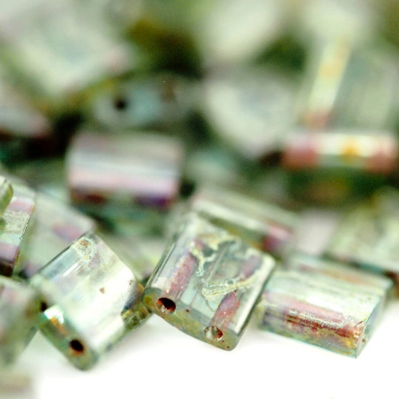 May include: A collection of rectangular glass beads featuring a translucent light green base with iridescent metallic finishes and internal streaks of reddish-brown and gold. Each bead has two small holes drilled through the center for threading.