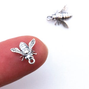 May include: Two small, silver-coloured bee charms. The detailed charms feature wings, body segments, and a loop for attaching to jewellery. One bee is in focus, resting on a fingertip, while another is slightly blurred in the background. The background is white.