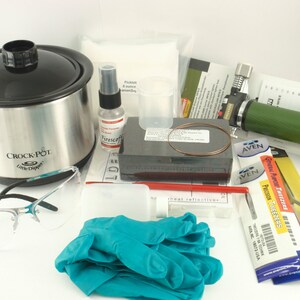 May include: A collection of jewelry-making tools and supplies, including a black and silver Crock-Pot, safety glasses, a green torch, precision tweezers, and blue gloves. Other items include a spray bottle, small containers, and various instruction sheets.
