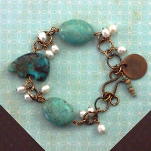 May include: A charm bracelet featuring two oval turquoise stones, a bear-shaped turquoise carving, and multiple white freshwater pearls. The components are connected by copper-toned metal rings and a hook clasp, with a round copper-toned metal disc.