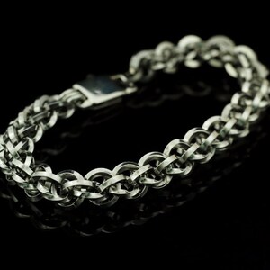 May include: A silver-toned chainmail bracelet constructed from interlocking circular metal rings. The bracelet features a repeating woven pattern and a rectangular metal clasp mechanism, showcasing a polished metallic finish throughout the design.