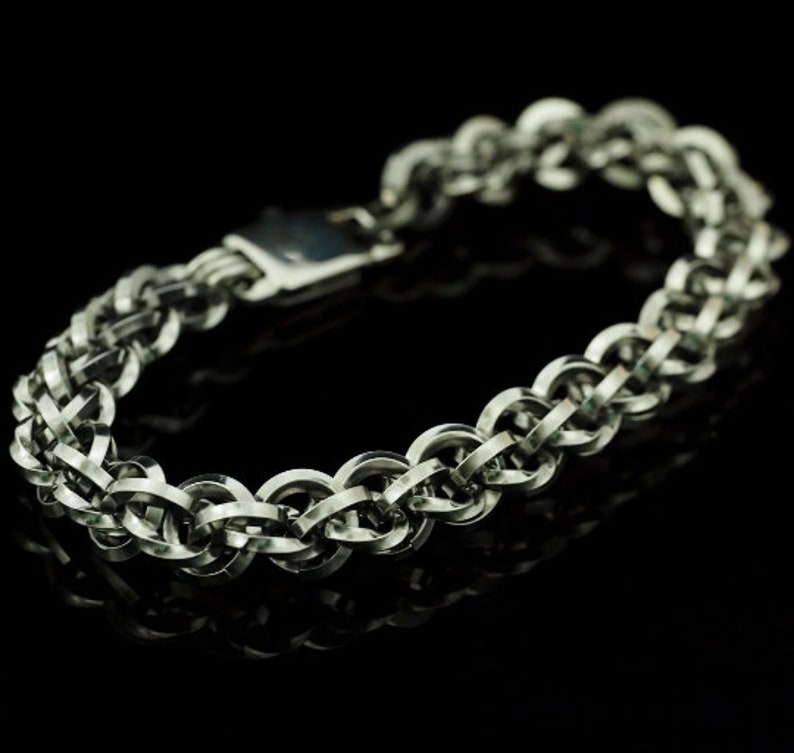 May include: A silver-toned chainmail bracelet constructed from interlocking circular metal rings. The bracelet features a repeating woven pattern and a rectangular metal clasp mechanism, showcasing a polished metallic finish throughout the design.