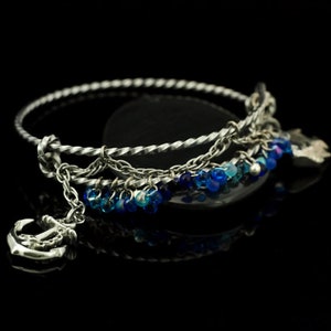 May include: A silver-toned metal bangle bracelet featuring a twisted band design. The bracelet is adorned with a dangling silver anchor charm and a series of small, translucent blue beads that hang from a delicate chain attached to the main band.