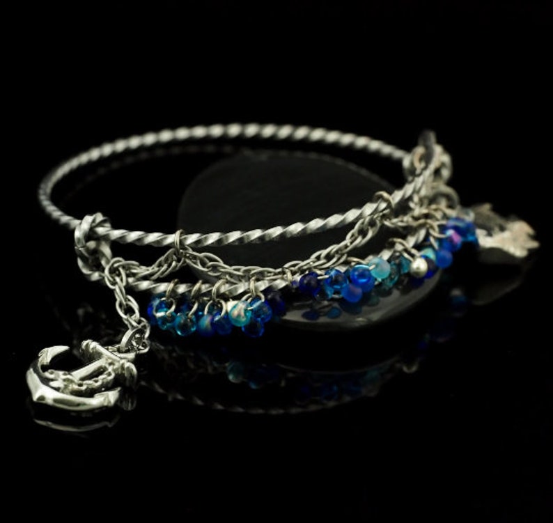 May include: A silver-toned metal bangle bracelet featuring a twisted band design. The bracelet is adorned with a dangling silver anchor charm and a series of small, translucent blue beads that hang from a delicate chain attached to the main band.