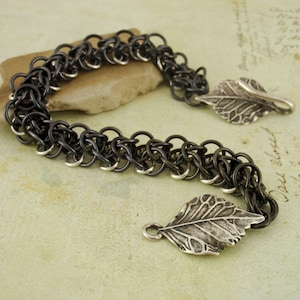 May include: A black chain bracelet with two silver leaf charms. The chain is made of small, interlocking rings.