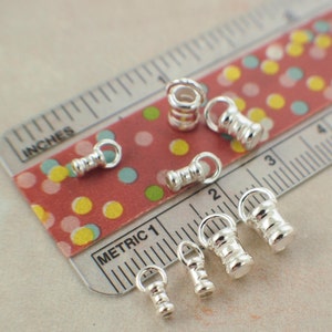 May include: A collection of small, shiny silver-colored metal jewelry findings, including barrel-shaped beads with attached loops. The findings are arranged on a red and white patterned surface next to a ruler marked in inches and metric units.