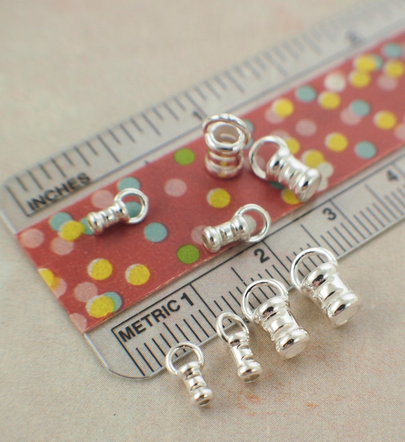 May include: A collection of small, shiny silver-colored metal jewelry findings, including barrel-shaped beads with attached loops. The findings are arranged on a red and white patterned surface next to a ruler marked in inches and metric units.