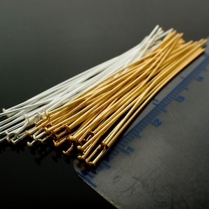 May include: Close-up of a collection of head pins in gold and silver tones. The pins are arranged in a neat pile, with a ruler in the background. The pins are used for jewelry making and crafting.