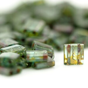 May include: A collection of small, rectangular glass beads featuring a translucent green and teal base with internal metallic gold and bronze-toned inclusions. Each bead has two parallel vertical holes running through the center of the glass.