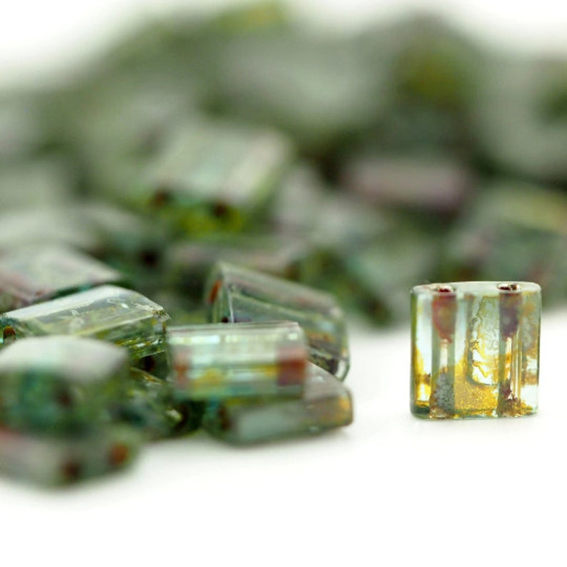 May include: A collection of small, rectangular glass beads featuring a translucent green and teal base with internal metallic gold and bronze-toned inclusions. Each bead has two parallel vertical holes running through the center of the glass.