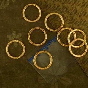 Puede incluir: Ocho anillos de salto de metal de color dorado, cada uno con una textura martillada. Los anillos est&aacute;n dispuestos sobre una superficie marr&oacute;n con una regla en el fondo.