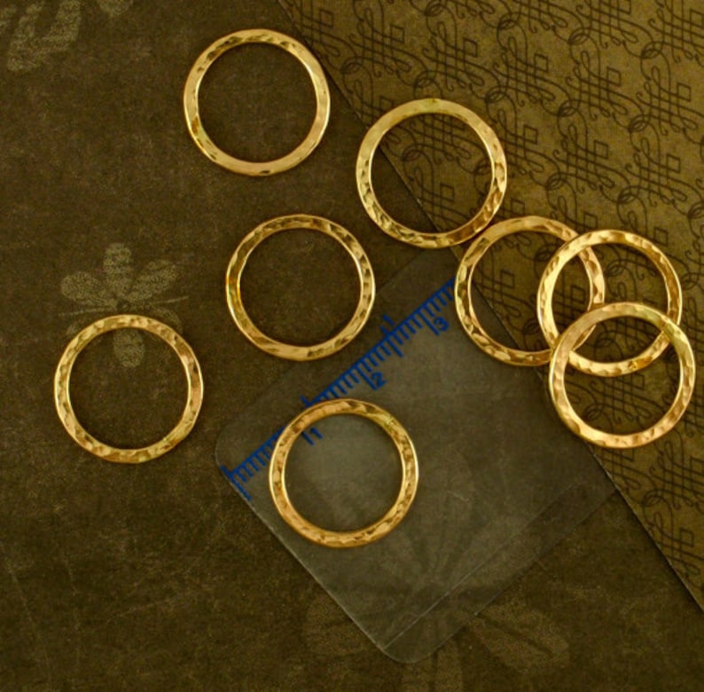 Puede incluir: Ocho anillos de salto de metal de color dorado, cada uno con una textura martillada. Los anillos est&aacute;n dispuestos sobre una superficie marr&oacute;n con una regla en el fondo.