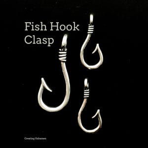 Op de afbeelding: Drie zilverkleurige metalen vishaaksluitingen van verschillende groottes, verticaal gerangschikt. Elke sluiting heeft een gebogen haakontwerp met een gewikkeld draaddetail aan de bovenste lus. De tekst "Fish Hook Clasp" en "Creating Unkamen" is zichtbaar.