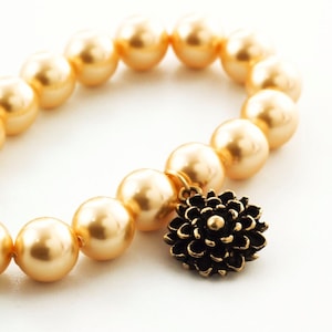 Puede incluir: Un brazalete dorado con grandes cuentas redondas de imitaci&oacute;n de perla y un dije de flor dorada.