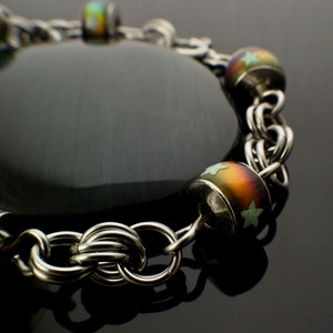 Puede incluir: Un brazalete de cadena de plata con una piedra negra y cuentas de vidrio de colores. Las cuentas tienen un diseño de estrella.