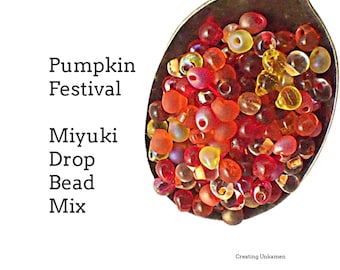 Puede incluir: Una cuchara de metal llena de una mezcla de pequeñas cuentas de vidrio en forma de gota en tonos rojos, naranjas y amarillos. El texto dice: Pumpkin Festival Miyuki Drop Bead Mix. Creating Unkamen aparece en la esquina inferior.