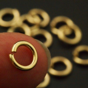 May include: A small, circular, gold-colored metal jump ring with a single open gap in the band. The jump ring has a smooth, polished surface and a reflective finish. Several additional identical gold-colored jump rings are scattered in the background.