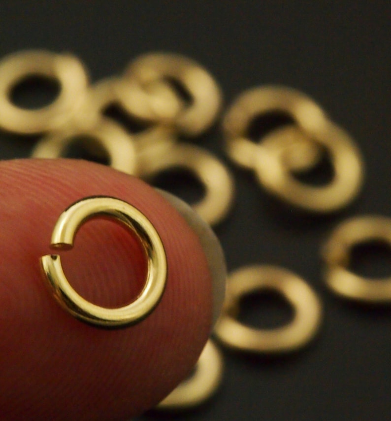 May include: A small, circular, gold-colored metal jump ring with a single open gap in the band. The jump ring has a smooth, polished surface and a reflective finish. Several additional identical gold-colored jump rings are scattered in the background.