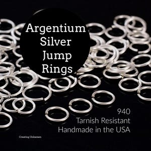 Op de afbeelding: Een verzameling zilverkleurige metalen springringen verspreid over een donker oppervlak. De tekst op de afbeelding luidt: Argentium Silver Jump Rings, 940 Tarnish Resistant Handmade in the USA, en Creating Unkamen. De ringen zijn cirkelvormig en open.