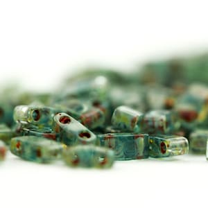 May include: A collection of small, rectangular glass beads in a translucent teal and blue hue. Each bead features two distinct holes and is decorated with subtle, swirling patterns of reddish-brown and yellow accents throughout the glass surface.
