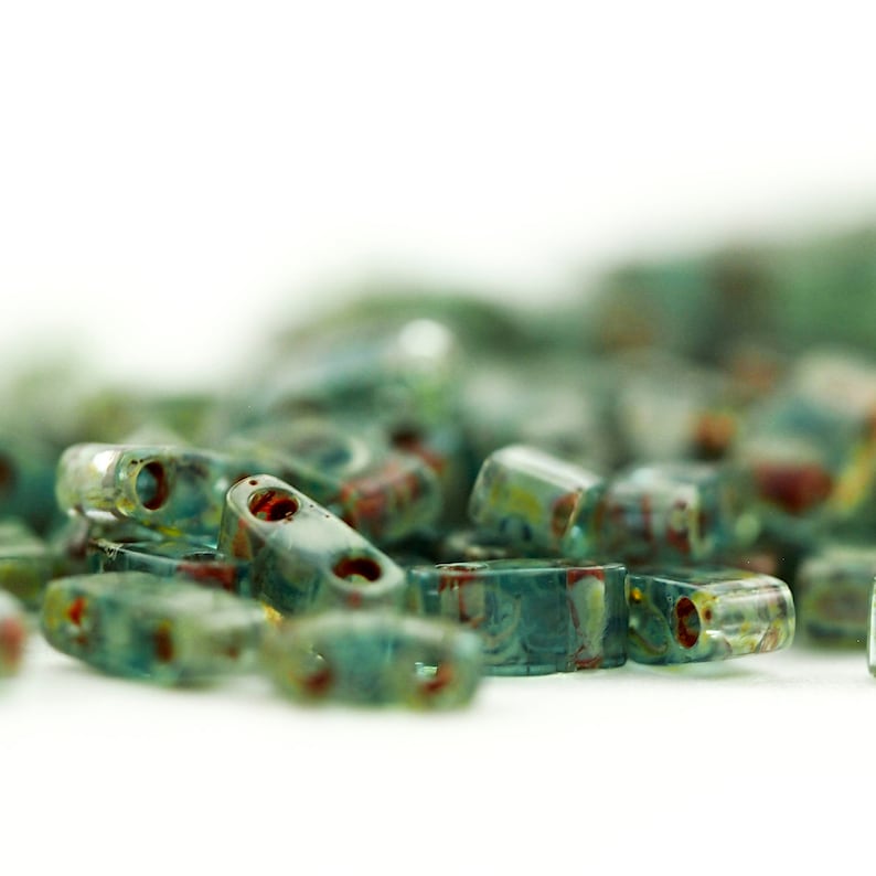 May include: A collection of small, rectangular glass beads in a translucent teal and blue hue. Each bead features two distinct holes and is decorated with subtle, swirling patterns of reddish-brown and yellow accents throughout the glass surface.