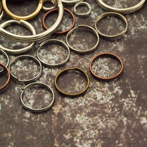 Op de afbeelding: Een verzameling verschillende metalen sleutelringen in verschillende maten en afwerkingen, waaronder zilver, goud en koper.