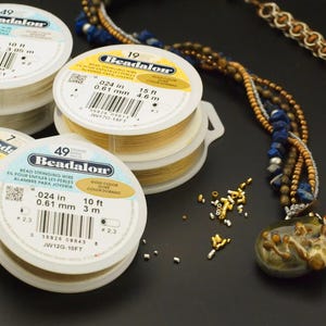 Puede incluir: Tres carretes de pl&aacute;stico blanco de hilo para abalorios Beadalon en colores oro y plata, con etiquetas que indican un di&aacute;metro de 0,61 mm y longitudes de 3 a 4,6 metros. Un collar de varias hebras con cuentas azules y marrones est&aacute; cerca.