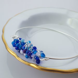 May include: A silver bangle bracelet featuring a cluster of blue, white and purple beads. The beads are arranged in a gradient pattern, with the darkest blue beads at the bottom and the lightest white beads at the top.