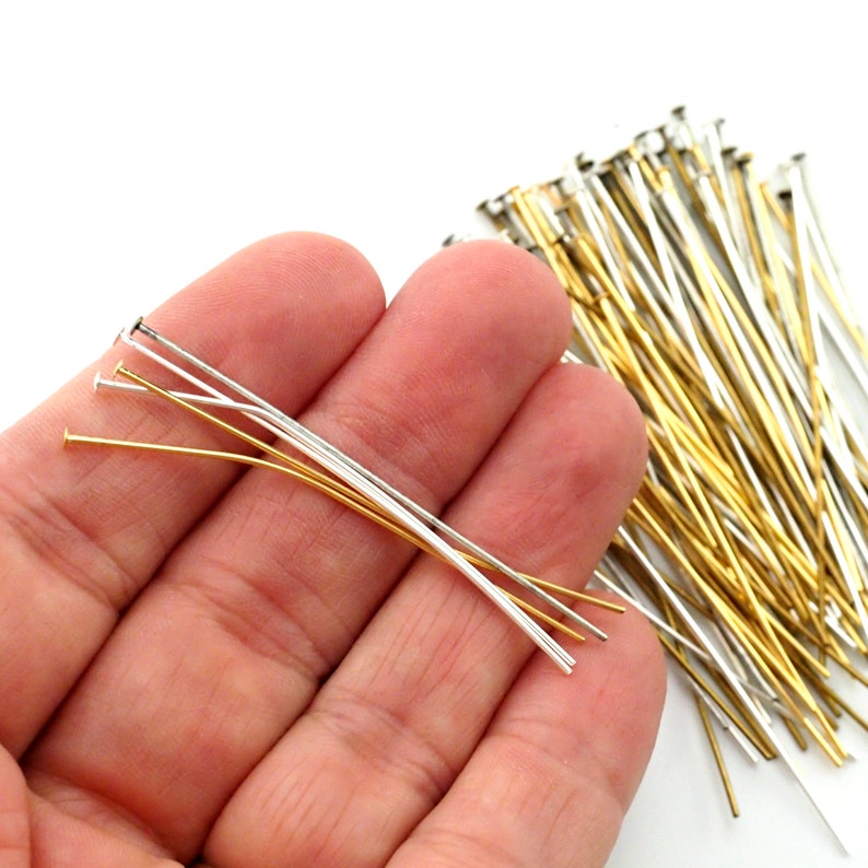 May include: A collection of head pins in gold and silver tones. The pins have a flat head and a long, slender body. Some pins are held in a hand, while the rest are scattered in the background. These pins are ideal for jewelry making and crafting.
