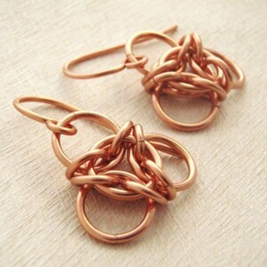 May include: A pair of copper earrings featuring a chainmaille design. Each earring is constructed from multiple interlocking circular copper rings, forming a triangular cluster that hangs from a curved copper ear wire hook for attachment.
