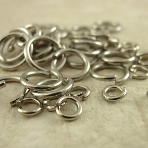Puede incluir: Una colecci&oacute;n de anillas de salto de metal plateado, cada una con forma circular y una abertura de corte &uacute;nico. Estos peque&ntilde;os accesorios circulares para joyer&iacute;a est&aacute;n hechos de un material met&aacute;lico pulido y reflectante, esparcidos en un mont&oacute;n.