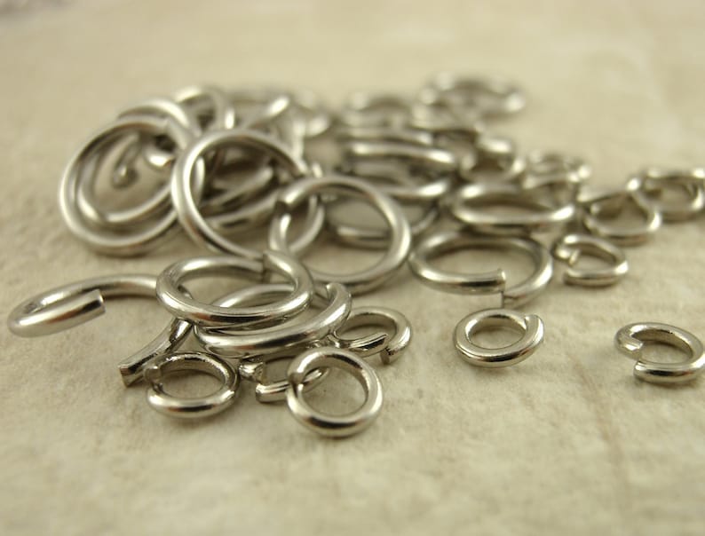 Puede incluir: Una colecci&oacute;n de anillas de salto de metal plateado, cada una con forma circular y una abertura de corte &uacute;nico. Estos peque&ntilde;os accesorios circulares para joyer&iacute;a est&aacute;n hechos de un material met&aacute;lico pulido y reflectante, esparcidos en un mont&oacute;n.