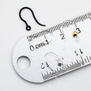 Puede incluir: Una colecci&oacute;n de ganchos para pendientes que incluye un gancho de metal negro mate y tres ganchos de pl&aacute;stico transparente, dos de los cuales tienen peque&ntilde;as cuentas de tono dorado y plateado. Una regla met&aacute;lica muestra una medida de 2 cm.