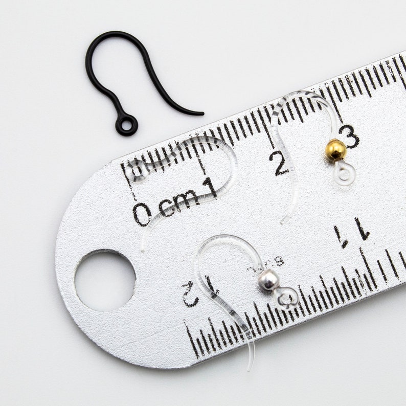 Puede incluir: Una colecci&oacute;n de ganchos para pendientes que incluye un gancho de metal negro mate y tres ganchos de pl&aacute;stico transparente, dos de los cuales tienen peque&ntilde;as cuentas de tono dorado y plateado. Una regla met&aacute;lica muestra una medida de 2 cm.