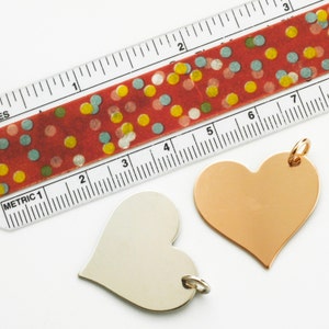 May include: Two heart-shaped metal tags, one silver and one gold, with a ruler in the background. The ruler has a red and white polka dot tape measure on top of it. The ruler is marked with "Metric 1" and numbers 1 through 7.