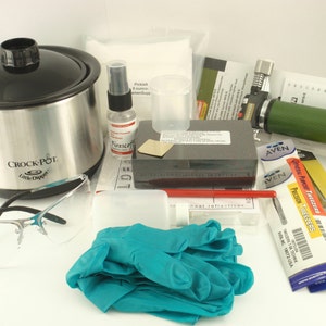以下が含まれることがあります： 小さなCrock-Pot、保護メガネ、トーチ、ピンセット、手袋、その他様々なアイテムを含む、ジュエリー作りのためのツールと用品のコレクション。白い表面に配置されています。