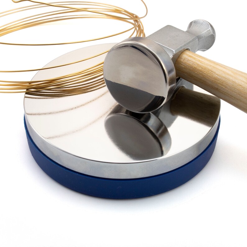 May include: A jewelry-making setup featuring a shiny, silver metal dapping block with a blue base, a metal hammer with a wooden handle, and coiled gold-colored wire. The hammer rests on the dapping block, suggesting a crafting process.