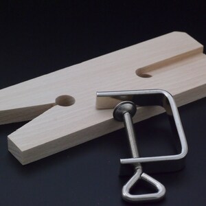 May include: A light-colored wooden bench pin with a V-shaped cutout and a central hole, accompanied by a silver-toned metal C-clamp featuring a threaded screw mechanism and a triangular handle for securing the tool to a workbench surface.