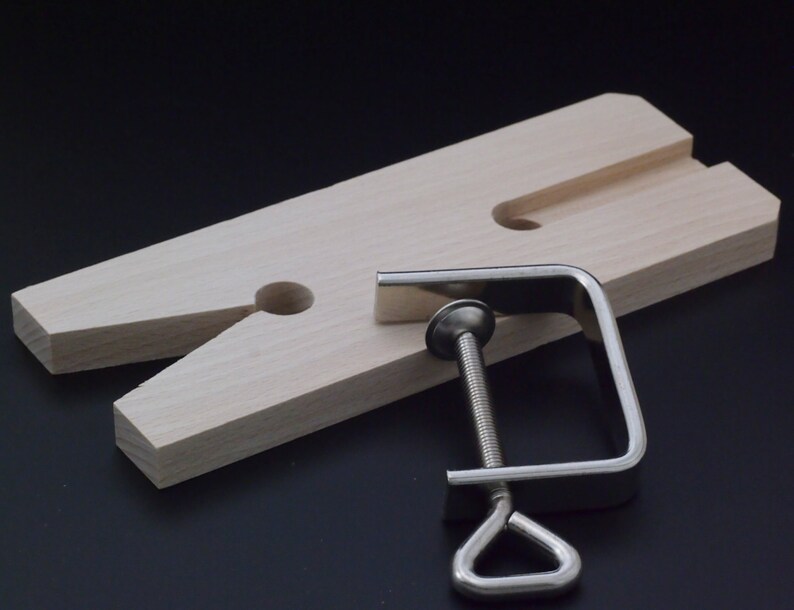 May include: A light-colored wooden bench pin with a V-shaped cutout and a central hole, accompanied by a silver-toned metal C-clamp featuring a threaded screw mechanism and a triangular handle for securing the tool to a workbench surface.