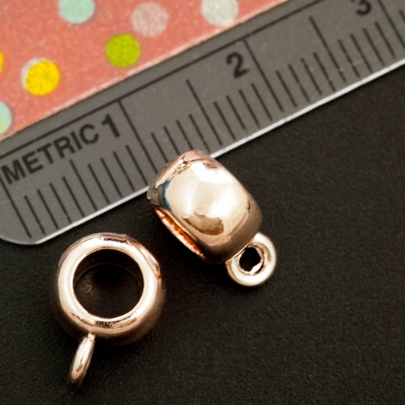 May include: Two rose gold metal bead caps with a round hole in the center and a small loop on the bottom. The caps are on a black surface.