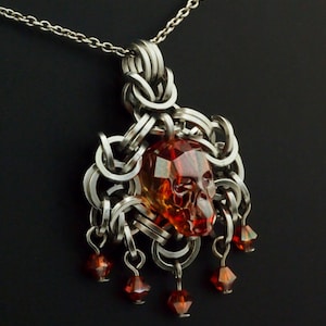 Op de afbeelding: Een zilveren ketting met een schedelvormige hanger van barnsteen en hangende rode kristallen kralen.