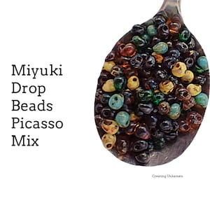 May include: A spoon filled with colourful Miyuki drop beads in a Picasso mix. The beads are a mix of brown, green, blue, and yellow.