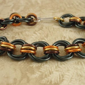 May include: A chainmaille bracelet constructed from interlocking rings in alternating colors of matte black, metallic copper, and bright gold. The circular links are woven together in a repeating pattern, secured by a simple silver-toned metal hook clasp.