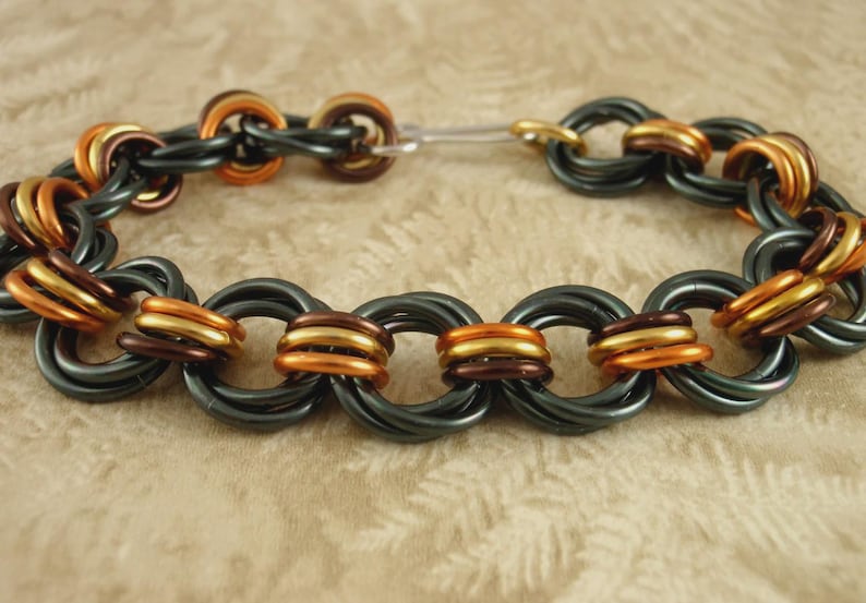 May include: A chainmaille bracelet constructed from interlocking rings in alternating colors of matte black, metallic copper, and bright gold. The circular links are woven together in a repeating pattern, secured by a simple silver-toned metal hook clasp.