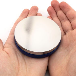 May include: A round, silver-colored object with a blue rim. The object is held in open hands against a white background. The top surface is reflective, and the blue rim adds a contrasting color.