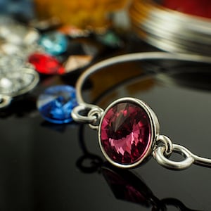 May include: A silver bangle bracelet featuring a large, faceted, pink gemstone. The bracelet is crafted from a thin, flexible metal and has a simple, elegant design.