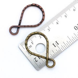 May include: Two metal jewelry findings, one is copper and the other is brass. The findings are shaped like a teardrop with a loop at the top. The findings are measured against a ruler.
