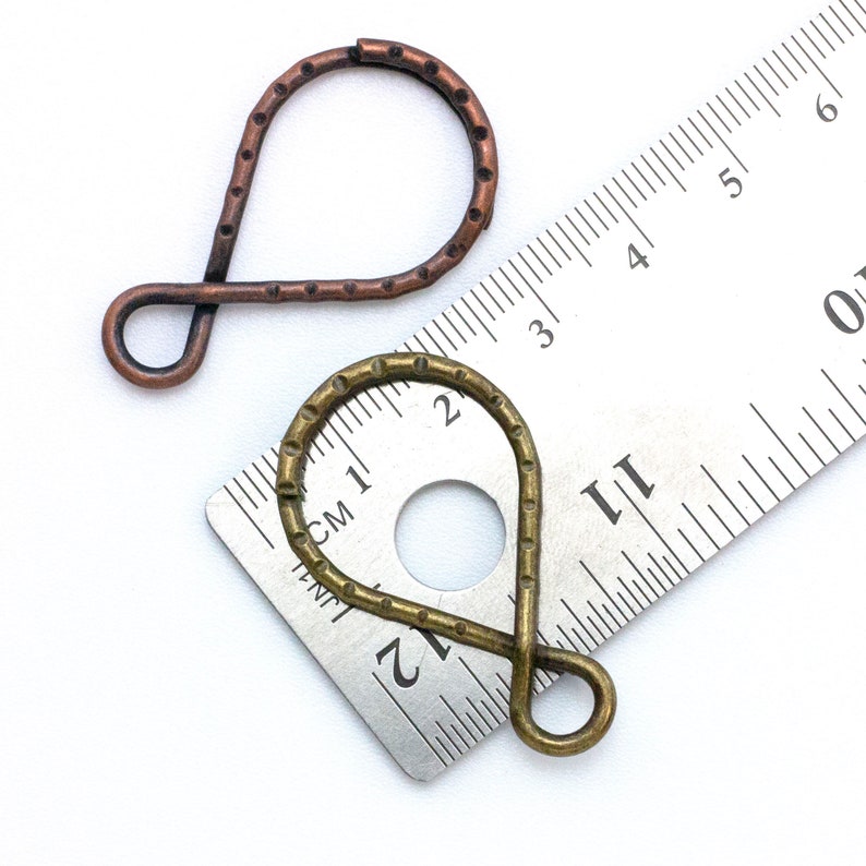 May include: Two metal jewelry findings, one is copper and the other is brass. The findings are shaped like a teardrop with a loop at the top. The findings are measured against a ruler.