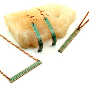 May include: A set of copper jewellery with a turquoise enamel finish. The set includes a necklace, a pair of earrings, and a pendant. The jewellery is displayed on a light brown stone.